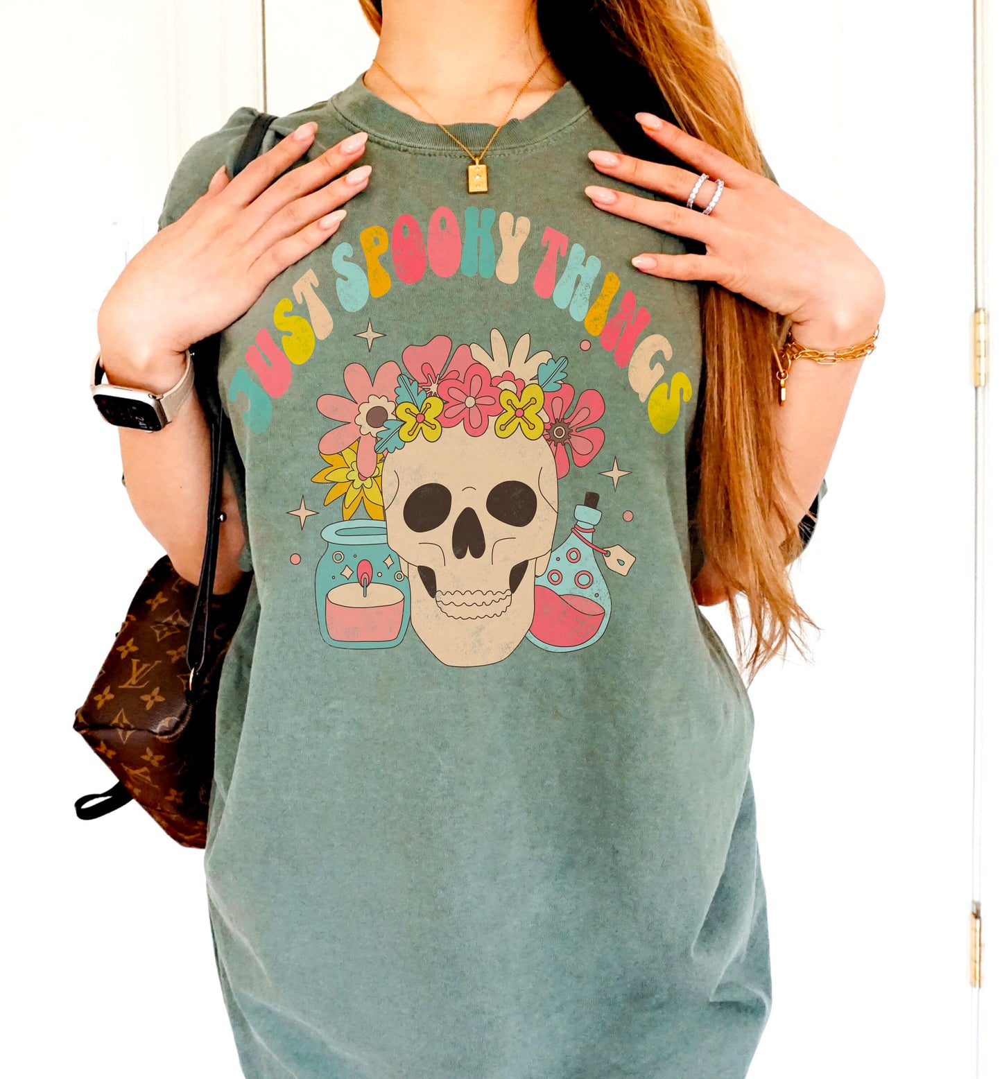 A t shirt in the color &quot;Moss&quot; with the words &quot;Just Spooky Things&quot; in Multicolor Retro Letters. Design features a skull with a flower crown, a candle, and a bottle of potion.