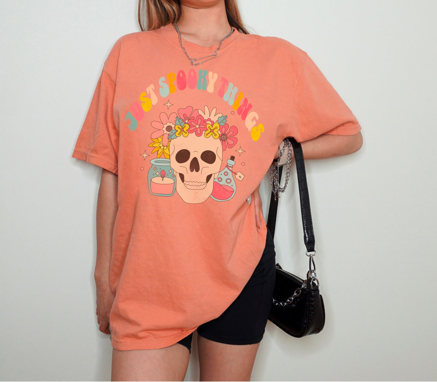 A t shirt in the color &quot;Terracotta&quot; with the words &quot;Just Spooky Things&quot; in Multicolor Retro Letters. Design features a skull with a flower crown, a candle, and a bottle of potion.